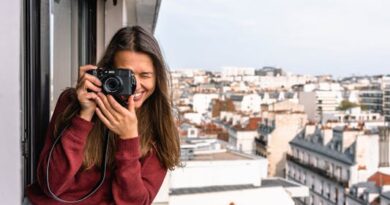 Woman Wearing Maroon Sweater Standing on Veranda Using Camera While Smiling Overlooking Houses and Buildings