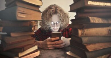 Focused male student in casual wear and glasses browsing smartphone while sitting at wooden table with stacked old books