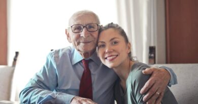 Positive senior man in formal wear and eyeglasses hugging with young lady while sitting at table